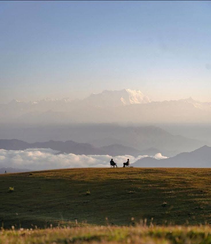 Two people sitting together on a hilltop, talking, overlooking mountains and clouds