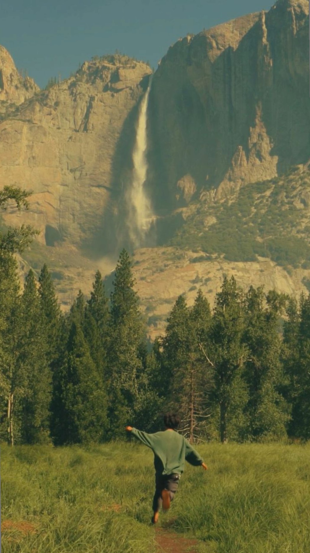 A person running through a meadow toward a waterfall and mountains